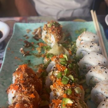 Dragon Sushi - A ceramic plate features an assortment of sushi rolls with various toppings and garnishes. Some rolls are topped with green onions, fish roe, and sesame seeds, while others have a drizzle of sauce. The plate is placed on a table with chopsticks resting on the side, and a person wearing a white T-shirt is partially visible in the background. - Sushi Bars Near Me - Albuquerque, New Mexico