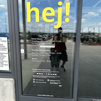 IKEA - A glass door at IKEA in Live Oak, TX, features large yellow text reading 'hej!' and additional information about store hours, Swedish restaurant hours, and payment options. The door reflects the parking lot, cars, and partly cloudy sky, with a person and child visible in the reflection. The signage provides details about the store's operation times and amenities. - Furniture Stores Near Me - Live Oak, Texas