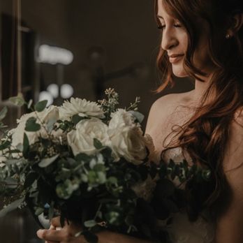 Process Salon - Wedding hair ! - A woman with long, wavy brown hair, wearing a strapless dress, is holding a bouquet of white roses and greenery. She is standing indoors near a mirror, with soft lighting highlighting her profile and the flowers. The background is blurred, emphasizing her and the bouquet. - Hair Salons Near Me - Phoenix, Arizona