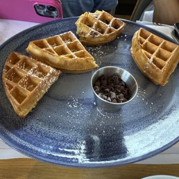 $18 Waffle for a child - Four waffle pieces, dusted with powdered sugar, are arranged on a large blue plate with a small container of chocolate curls in the center.