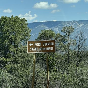 Fort Stanton Museum - A brown directional road sign with white text points left towards Fort Stanton State Monument, situated among green trees with a mountainous landscape in the background. The sky is partly cloudy with scattered clouds, and the sign is supported by two metal posts. The image appears to be taken from a vehicle, with part of the car's dashboard visible at the bottom. - Museums Near Me - Fort Stanton, New Mexico