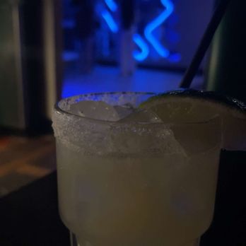 Night Train - NT Marg - A drink with ice and a lime wedge in a glass is placed on a dark surface at a bar, with blue neon lighting in the background. - Cocktail Bars Near Me - Fort Wayne, Indiana