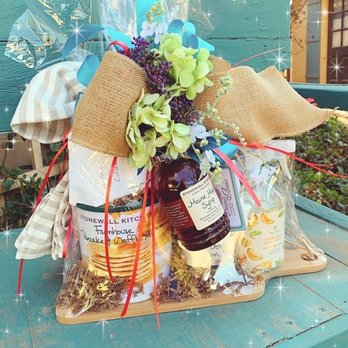 Bonnes Amies - The best gift wrapping in Plainview! Buy something and they'll kindly gift wrap if needed! - A gift basket is displayed on a wooden surface, wrapped in clear plastic with red ribbons. It contains a bouquet of flowers with purple, green, and white blooms, a jar of Maine Maple Syrup, and food items including pancake and waffle mix and a can of mandarin oranges. The basket is decorated with burlap and fabric ribbons, and the background features a teal wooden surface with sparkles added for effect. - Interior Design Near Me - Plainview, Texas