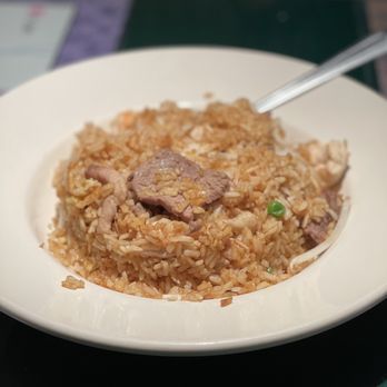 China Cafe - Combination Fried Rice - A white bowl contains fried rice with small pieces of meat and green peas, placed on a dark surface. A spoon is partially inserted into the rice, and the background is blurred, showing a table setting and some indistinct objects. - Chinese Near Me - Durango, Colorado