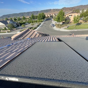 Clear Choice Cleaning - The image captures a high-angle view from a rooftop, showing a residential neighborhood with houses, trees, and a curved street with parked cars. The background features mountain ranges under a partly cloudy blue sky. The rooftop includes roofing materials and solar panels, indicating ongoing or completed roof work. The scene is calm and sunny, with well-maintained lawns and a scenic mountain backdrop. - Solar Panel Cleaning Near Me