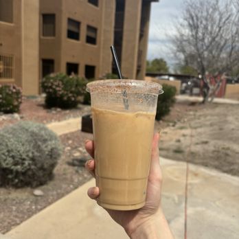 Roadrunner Coffee - Iced latte - A person’s hand, wearing a yellow sleeve, holds a clear plastic cup filled with iced coffee and a black straw. The background features a multi-story building with a beige exterior, some bushes, and leafless trees, indicating an outdoor setting on a cloudy day. - Coffee & Tea Near Me - Tucson, Arizona