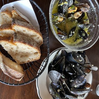 The Dock - Out of this world mussels ! - The image shows a plate of cooked mussels, a bowl of mussels in broth with garlic and herbs, and a basket of toasted bread slices on a dark wooden table. - Seafood Near Me - Montauk, New York