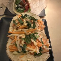 Falafel Wrap - Three tacos filled with shredded carrots, cabbage, and herbs are arranged on a black plate. The tacos are topped with a creamy sauce and fresh herbs. In the background, there is a taco and a wrap on a separate plate, partially wrapped in paper. The setting appears to be a casual dining environment.