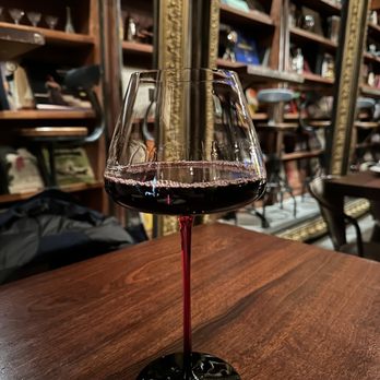 Tavern 157 - A glass of red wine is placed on a wooden table inside a wine shop or bar, with shelves of bottles and decorative items in the background. - Lounges Near Me - Flushing, New York