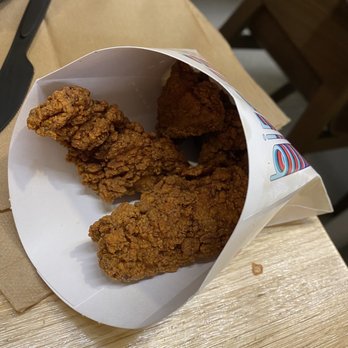 Birdcall - All Natural Chicken - Three pieces of fried chicken with crispy, golden-brown coating are placed inside a white paper container on a light-colored wooden table. The container is slightly crumpled at the edges, and a pair of black tongs is positioned nearby on the table. The background includes a cardboard piece and part of a chair, indicating an indoor setting, likely a casual dining or takeout environment. - Chicken Shop Near Me - Denver, Colorado