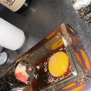 Nohri - Beef tartare - A sushi roll with rice, fish, and seaweed is in a glass container, alongside a raw egg yolk in soy sauce, on a dark countertop. - Sushi Bars Near Me - Boston, Massachusetts