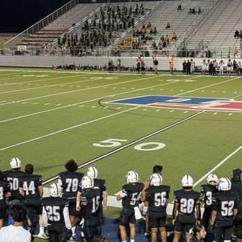 The Pfield - Football players in black uniforms with white helmets are seated on the sideline of a football field, facing the field. The field has yard lines and a logo at the center. Spectators and team staff are visible near the sideline, with some standing and others sitting. The stadium has bleachers with some spectators seated, and the scene is illuminated by stadium lights at night. - Stadiums & Arenas Near Me - Pflugerville, Texas