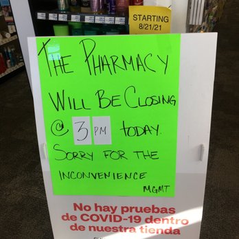 CVS Pharmacy - Sign inside the store - The image shows a bright green handwritten sign placed on a white stand inside a store, likely a pharmacy. The sign states that the pharmacy will close today at 3 PM and apologizes for the inconvenience. It also includes a notice in Spanish indicating that no COVID-19 tests are available inside the store and provides instructions for appointment scheduling. The background features shelves with personal care products and a yellow note indicating a start date of 8/21/21. - Drugstores Near Me - Goshen, Indiana