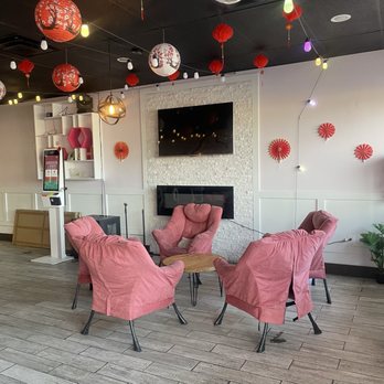 KUTEA - The image shows four pink upholstered armchairs arranged around a small round wooden table in an indoor setting. The ceiling is decorated with hanging lanterns and string lights, some of which are shaped like trees and flowers. The wall behind the seating area features a mounted flat-screen TV, a white textured wall panel, and decorative red wall ornaments. The floor is covered with wood-look tiles, and there is a potted plant on the right side of the image. - Desserts Near Me - Denver, Colorado