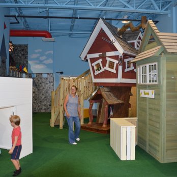 Unplug and Play - Indoor playground with themed playhouses, including a red and white house and a green house, along with a wooden climbing structure and slide. A woman stands nearby, supervising children, with a child playing and another sitting inside a playhouse. The environment has artificial green turf flooring and colorful wall decorations, creating a safe and engaging space for children. - Indoor Playcenter Near Me - Montclair, Virginia