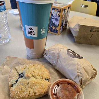 Satellite Coffee - Early bird burrito, blueberry scone and Dirty Chai Latte - A coffee cup, blueberry scone, and wrapped sandwich are placed on a table at Satellite Coffee, with a water bottle and decorative box in the background. - Coffee & Tea Near Me - Albuquerque, New Mexico
