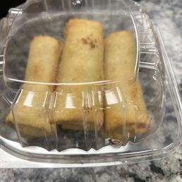 Philly Cheesesteak Egg Rolls - A clear plastic container holds five egg rolls, arranged in a row, on a gray marble table. The container's lid is closed, and the egg rolls are golden-brown with some darker spots. The background is blurred, showing part of a chair and a dark area, indicating an indoor or outdoor dining setting.
