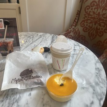 My Brigadeiro - Passion fruit mousse, mocha, chocolate croissant - The image shows a marble table with a small cup of mango or passion fruit mousse, a disposable coffee cup with a lid, and a plastic bag with the My Brigadeiro logo. There are also a pair of earphones, a key fob, and a small clear plastic container on the table. The background includes a patterned upholstered chair and a partial view of the restaurant interior. - Chocolatiers & Shops Near Me - Hanover, New Hampshire