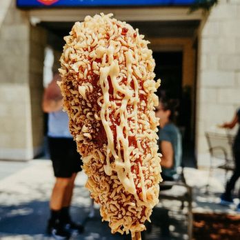 Mochi Ring - The half mozzarella half dog ramen - A hand holds a mochi donut on a stick, coated with crunchy toppings and drizzled with sauce, in front of the Mochi Ring storefront. The shop offers mochi donuts and Korean corn dogs, with a sign displaying the business name and logo. The background includes a sidewalk, trees, and a few people near the entrance, indicating an outdoor setting. - Donuts Near Me - Wauwatosa, Wisconsin