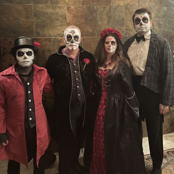 A Face Painter - Four individuals in Día de los Muertos costumes with skull face paint stand against a stone wall, wearing elaborate outfits and accessories. - Face Painting Near Me
