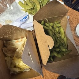 The image shows two cardboard takeout containers on a wooden table, one filled with cooked edamame beans and the other partially open, revealing a dish with melted cheese and possibly vegetables. A paper towel with additional edamame beans is placed nearby, along with a black mesh container and a digital scale. The setting appears to be a casual dining or takeout environment, with food items and utensils arranged on the table.