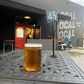 Chama Local - Patio People!!! - A clear glass of beer is positioned on a metal outdoor table with a textured surface. In the background, there is a black building with white text reading 'LOCAL' repeated multiple times and a red door. Two people are seated outside, one wearing a blue jacket and a hat, with a backpack nearby. A small sign with the number 45 is attached to a stand on the table, indicating a table number. The setting appears to be an outdoor dining area of a restaurant or cafe. - Pizza Near Me - Chama, New Mexico