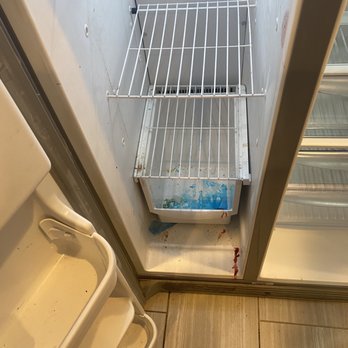 RentProv Realty - Our "clean" fridge on move in day. - The image depicts the interior of an open refrigerator with two wire shelves, both of which are empty. The bottom of the refrigerator has visible dirt and stains, including blue and red marks. The surrounding area includes a tiled floor and part of a plastic or metal shelf unit nearby. The refrigerator appears to be unclean and unused. - Real Estate Services Near Me - Providence, Rhode Island