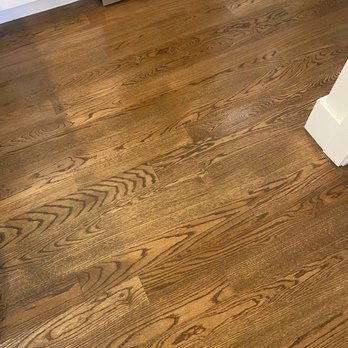 Dream House Services - New and old flooring - The image depicts a section of a kitchen floor made of hardwood with a warm, natural finish. The floorboards display visible wood grain patterns and are arranged in a parallel pattern. In the background, part of a white kitchen cabinet and a stainless steel oven are visible, indicating a modern kitchen setting. - General Contractors Near Me - Randolph, Massachusetts