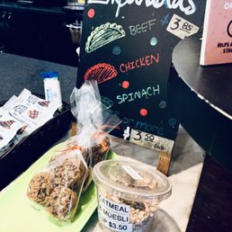 Some Bites - The image shows a chalkboard menu with the word 'Empanadas' at the top, listing fillings like beef, chicken, and spinach, along with prices. In front of the menu, there is a green tray holding packaged cookies and a clear plastic container labeled 'Oatmeal & Muesli Cup $3.50,' which contains a mixture of oats and dried fruits or nuts. The background includes a counter with various items, indicating a food stand or cafe setting.