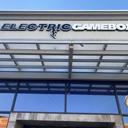 The photo captures the front of a commercial building with a sign displaying 'ELECTRIC GAMEBOX' in bold, stylized letters. The building features large glass windows and a modern architectural design, with a clear blue sky overhead.