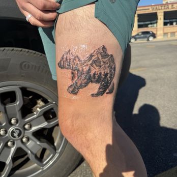 Sido Tattoo Studio - The tattoo features a mountain landscape with detailed peaks and forested areas, integrated into the shape of a bear. It is located on the upper thigh of a person wearing teal shorts, with sunlight illuminating the skin and tattoo. The background includes a parking lot with a vehicle and a building in the distance. - Tattoo Near Me - Chicago, Illinois
