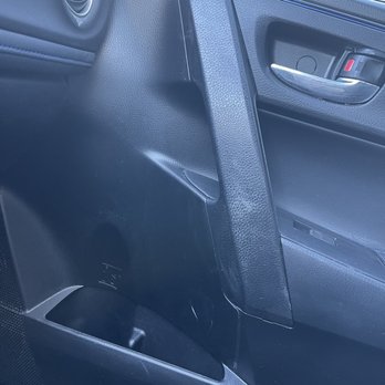 ProntoWash - A detailed view of a car's interior focusing on the passenger side door panel and dashboard. The door panel features a handle with a lock/unlock switch, and the dashboard includes a circular air vent. The materials are primarily black plastic with textured surfaces, and part of the windshield is visible at the top of the image. - Car Wash Near Me - Brandon, Florida