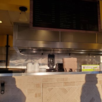 Gary Burritos - his shadow on the counter - Counter area in a restaurant with disposable food containers stacked on the counter, a cash register, and a menu board hanging above. The background includes a stainless steel surface and a brick wall below the counter. Shadows of people are visible on the brick wall. - Mexican Near Me - Carol Stream, Illinois