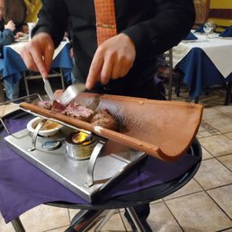 Filet Mignon - A waiter slices grilled meat on a wooden board at a restaurant table, with diners and table settings visible in the background.
