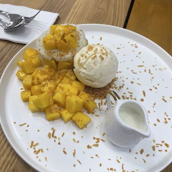 Mango Mango Dessert - A white round plate holds a serving of diced mango, a scoop of vanilla ice cream, and a pastry topped with coconut shavings and mango pieces. A small white pitcher of coconut milk is also on the plate. The plate is on a wooden table with a napkin and spoon nearby. - Desserts Near Me - Plainview, New York
