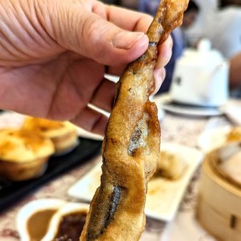 Fortune Villa - Salted fried capelin 多春魚 - A person holds a fried salted fish fillet, with a restaurant table setting in the background, including dipping sauces and Asian dishes. - Chinese Near Me - Markham, Ontario