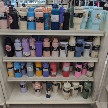 Winners Apparel - Drinkware - The image shows a retail store shelf filled with an assortment of reusable water bottles in multiple colors, including pink, purple, blue, black, and green. The bottles are arranged on four shelves, with some featuring straws and handles. The background reveals store aisles, shelves with clothing and accessories, and a customer browsing. The store appears well-lit and organized, with signage indicating discounts and product categories. - Women's Clothing Near Me - Gloucester, Ontario