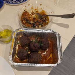Muhammara - A foil tray contains five meatballs in tomato sauce, accompanied by a bowl of herb and spice sauce with a spoon, on a white table.