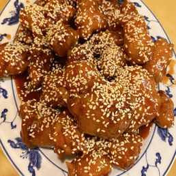Sesame Chicken - A serving of sesame chicken is presented on a blue and white patterned oval plate. The chicken pieces are coated in a thick, glossy sauce and topped with sesame seeds. The plate is placed on a wooden surface, and the dish appears to be a Chinese cuisine offering, featuring large portions suitable for sharing or leftovers.