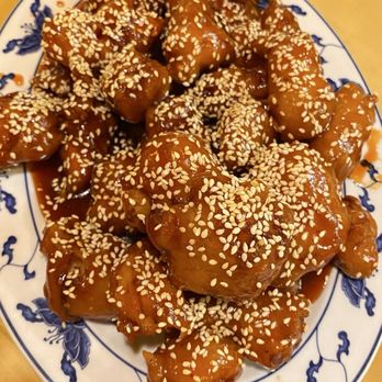 Royal Dragon - Sesame Chicken - A serving of sesame chicken is presented on a blue and white patterned oval plate. The chicken pieces are coated in a thick, glossy sauce and topped with sesame seeds. The plate is placed on a wooden surface, and the dish appears to be a Chinese cuisine offering, featuring large portions suitable for sharing or leftovers. - Chinese Near Me - Roy, Utah
