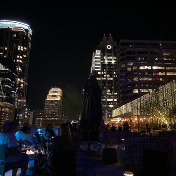 Edge Rooftop - great view - Night scene at a rooftop bar featuring city skyscrapers with illuminated windows and modern architecture. The bar area has string lights along the railing, creating a festive atmosphere. Several people are seated and standing, engaging in conversations, with some using their phones. The skyline includes tall buildings with bright lights, and the overall setting suggests a social gathering in an urban environment. - Lounges Near Me - Austin, Texas
