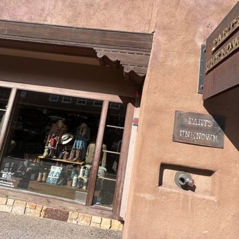 Parts Unknown - @ Santa Fe Plaza - Exterior view of a retail store with a large display window showing clothing and accessories, and signs reading "Parts Unknown" on the wall. - Shoe Stores Near Me - Santa Fe, New Mexico