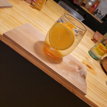 Soule' Coffee + Bubble tea - Peach Spritzer - A beverage called a peach spritzer, containing lemon slices and blueberries, is presented in a clear glass on a wooden serving board. The background shows a kitchen or cafe environment with various bottles, containers, and a person working at a counter. The setting suggests a casual dining or beverage preparation area. - Coffee & Tea Near Me - Jackson, Mississippi