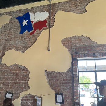 Alpha Omega Grill & Bakery - Brick stucco interior wall - The image shows an interior brick wall with sections of peeling beige paint, exposing the brick beneath. A Texas flag artwork is mounted on the wall, and a window with a black frame reveals parked cars and outdoor scenery. There are people seated at tables, and a string with framed pictures or notes is hung across the wall. - Coffee & Tea Near Me - Waco, Texas