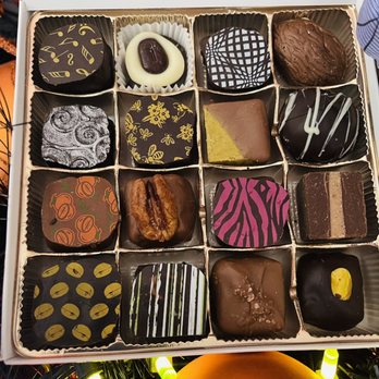 Serenade Chocolatier - Absolutely delicious! - A square box of artisan chocolates features 16 pieces arranged in a 4x4 grid. Each chocolate has unique decorations, including patterns, drizzles, and toppings such as nuts and colored coatings. The chocolates are crafted with different flavors and fillings, reflecting European-style confectionery. The box is open, revealing the chocolates' detailed designs and textures, and is set against a background with festive lighting. - Chocolatiers & Shops Near Me - Brookline, Massachusetts