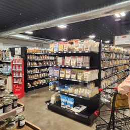 a view of a grocery store - The image shows an aisle in a grocery store with black metal shelves stocked with various packaged food items such as grains, pasta, and baking ingredients. The ceiling has exposed ductwork and bright overhead lighting. A shopping cart is visible on the right side, and a small display with jars is in the foreground. The store appears organized and well-lit, with a sign indicating frozen products in the background.