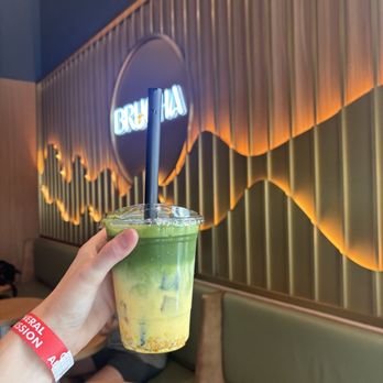 Brucha - Mango matcha - A person’s hand, wearing a red wristband, holds a clear plastic cup filled with a layered green and yellow drink, topped with a black straw. The cafe interior features a decorative wall with vertical wooden slats, warm lighting, and a neon sign that reads 'BRUCHIA'. The seating includes green cushioned benches and wooden tables, creating a contemporary atmosphere. - Gelato Near Me - Tucson, Arizona