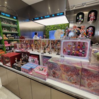 Pop Mart - The image shows a display of collectible figurines and toys at Pop Mart in Oak Brook, Illinois. The display includes multiple glass cases filled with anime-style figures, some in colorful packaging labeled 'Peach Peach' and 'Rainbow Kid.' Behind the display, there are shelves with additional merchandise, and a few customers are seen interacting at the counter. The store features a modern interior with metallic accents and bright lighting, emphasizing the variety of collectibles available for purchase. - Toy Stores Near Me - Oak Brook, Illinois