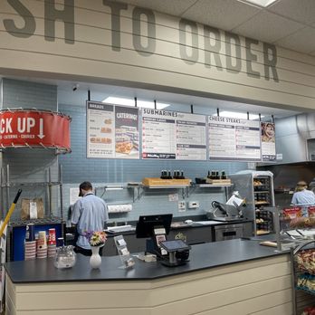 Jersey Mike's Subs - Inside Jersey Mike's Subs, a counter with menu boards overhead, staff preparing sandwiches, and various supplies are visible. - Fast Food Near Me - Westerly, Rhode Island