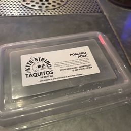 a plastic container of taquios - A clear plastic container labeled 'Kite String Taquitos' with Poblano pork taquitos inside, placed on a metal surface near a drain.